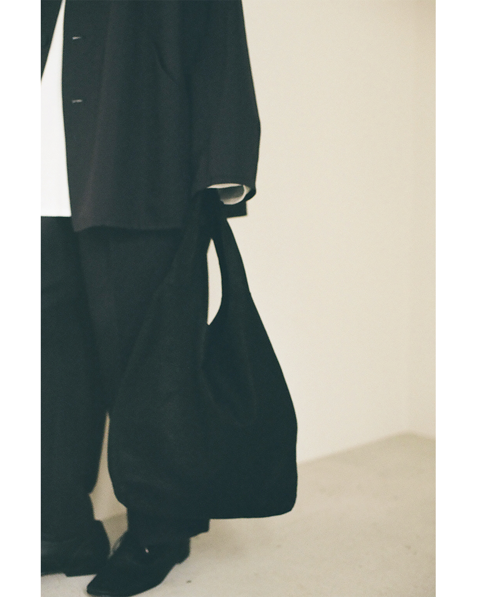 VUm（ヴウエム）の折りたためる黒の鹿革トートバッグ（Deer Leather Packable Tote black B23）を手で持った、ゆったりとしたユニセックスな黒セットアップのスタイリングカット